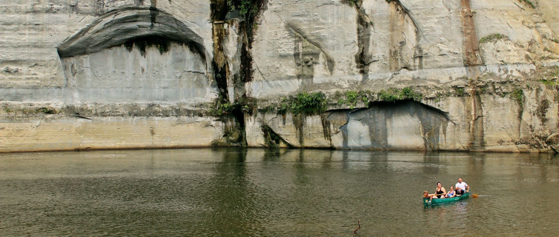 Group of people paddling a canoe on a calm river in front of a large rock wall with carved and weathered features.